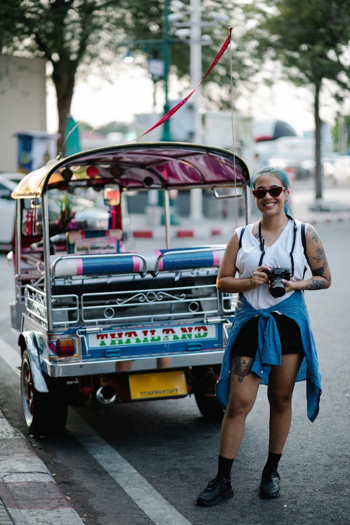 Young woman with camera beside colorful tuk-tuk on a bustling city street.